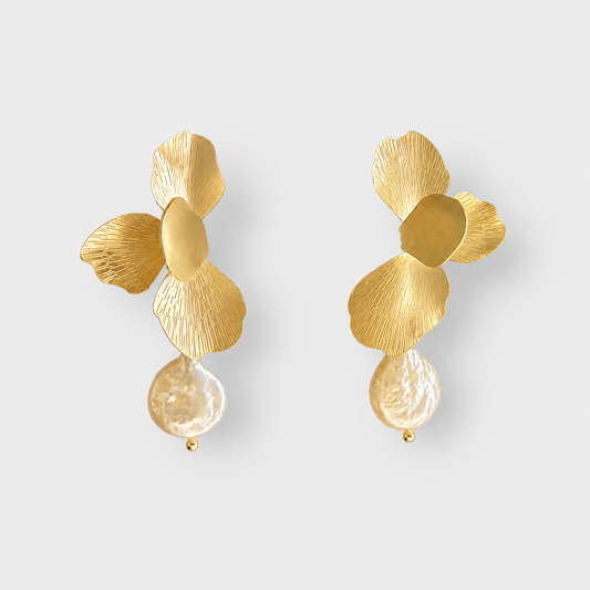 A pair of gold flower-shaped drop earrings with textured petals and a clear teardrop-shaped bead hanging below, shown on a light gray background.